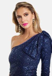 Marineblå glitterkjole med en enkelt off-the-shoulder design og en stor sløjfepynt. Skinnende tekstur og tætsiddende form.