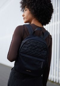 Mochila negra de nylon acolchado con una parte superior redondeada, correas lisas y un bolsillo frontal; logo de la marca en dorado visible en la parte inferior.