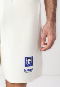 Κοντινό πλάνο ενός ατόμου που φοράει εκρού αθλητικά σορτς με μπλε λογότυπο Hummel Sportswear κοντά στο τελείωμα, στέκεται με χαλαρό χέρι.