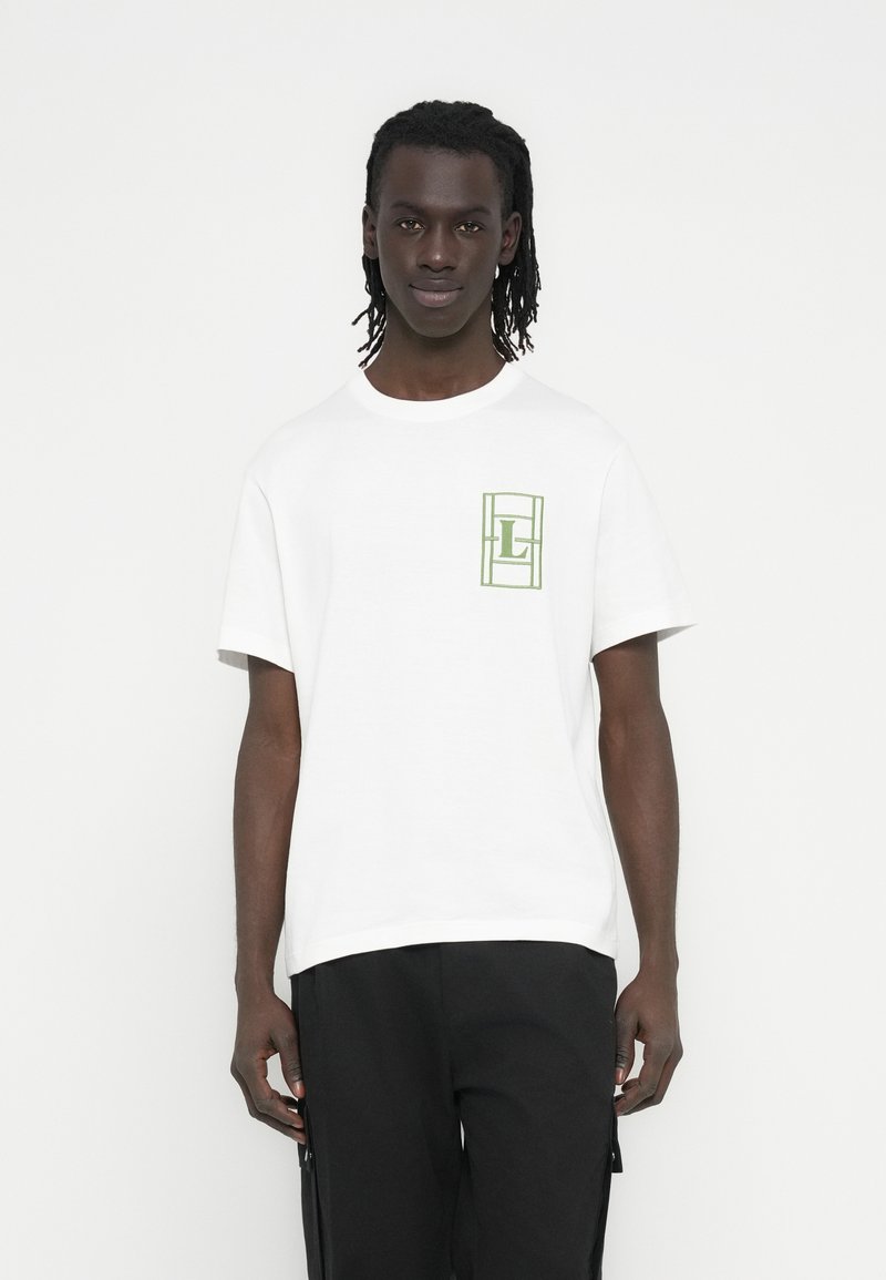 Λευκό βαμβακερό μπλουζάκι με κοντά μανίκια, που φέρει ένα πράσινο γεωμετρικό σχέδιο στη αριστερή πλευρά. Το μοντέλο φοράει μαύρο παντελόνι.