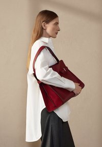 Bolso de cuero rojo con un acabado liso, forma estructurada y un acento de hardware plateado. Llevado sobre una camisa blanca y pantalones grises.