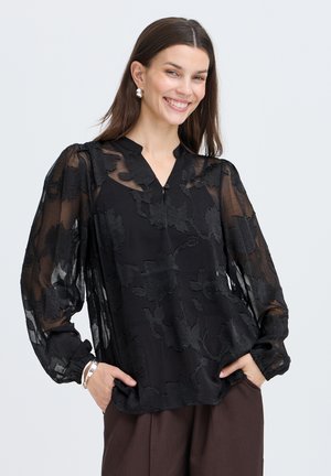 Femme souriante portant un chemisier noir transparent à motifs floraux avec des manches longues bouffantes et un pantalon marron foncé, mains dans les poches, sur un fond uni.