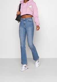 Osoba v růžové zkrácené mikině Hello Kitty, modrých džínách s vysokým pasem, bílých teniskách a s malou černou kabelkou přes rameno.