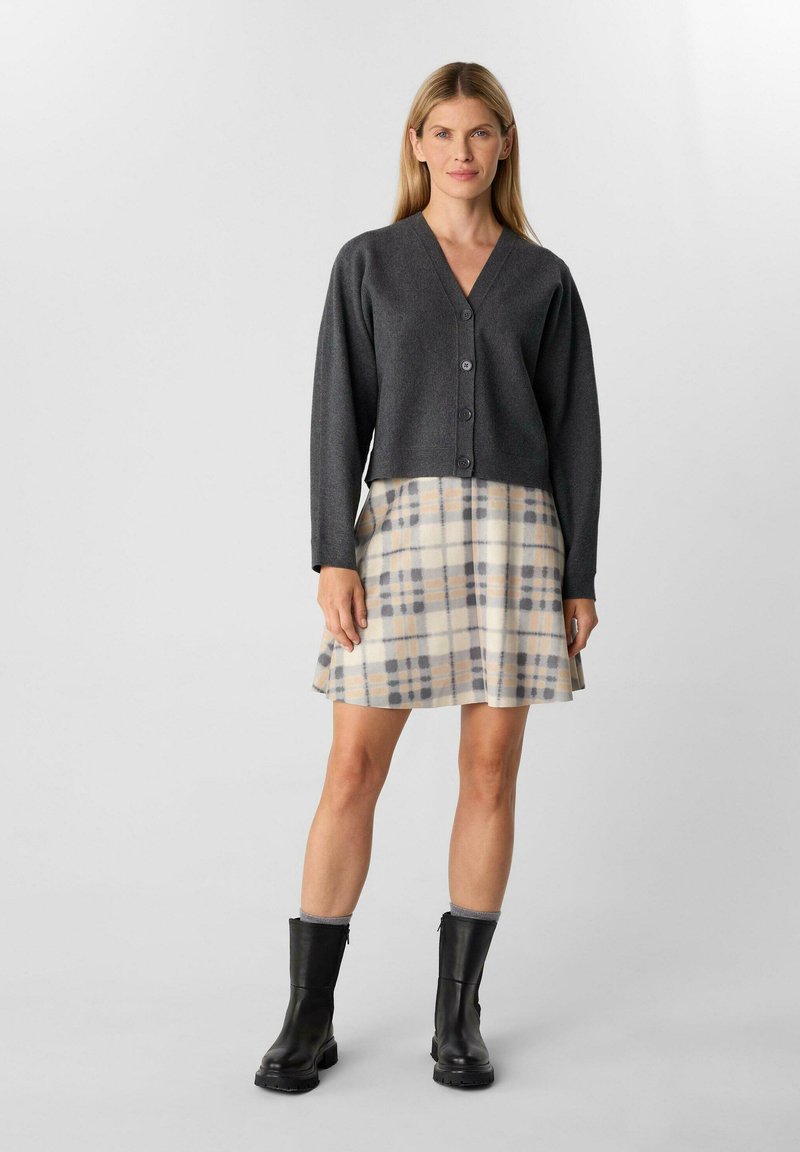 Cardigan grigio corto con scollatura a V e chiusura a bottoni, abbinato a una gonna a quadri crema con accenti blu e arancioni. Stivaletti neri alla caviglia.