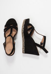 Černé semišové sandály na klínu s křížovým designem popruhů, s přezkou na kotníku a tkanými provazovými akcenty podél základny. Hladká vnitřní podšívka.