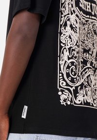 La camiseta de algodón negra presenta un diseño gráfico blanco con patrones intrincados. Incluye una pequeña etiqueta de logo en el lado inferior.