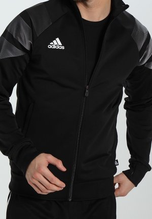 Homme portant une veste zippée noire Adidas avec un logo blanc et des rayures grises à motifs sur les épaules, mains près de la fermeture éclair et de la hanche.