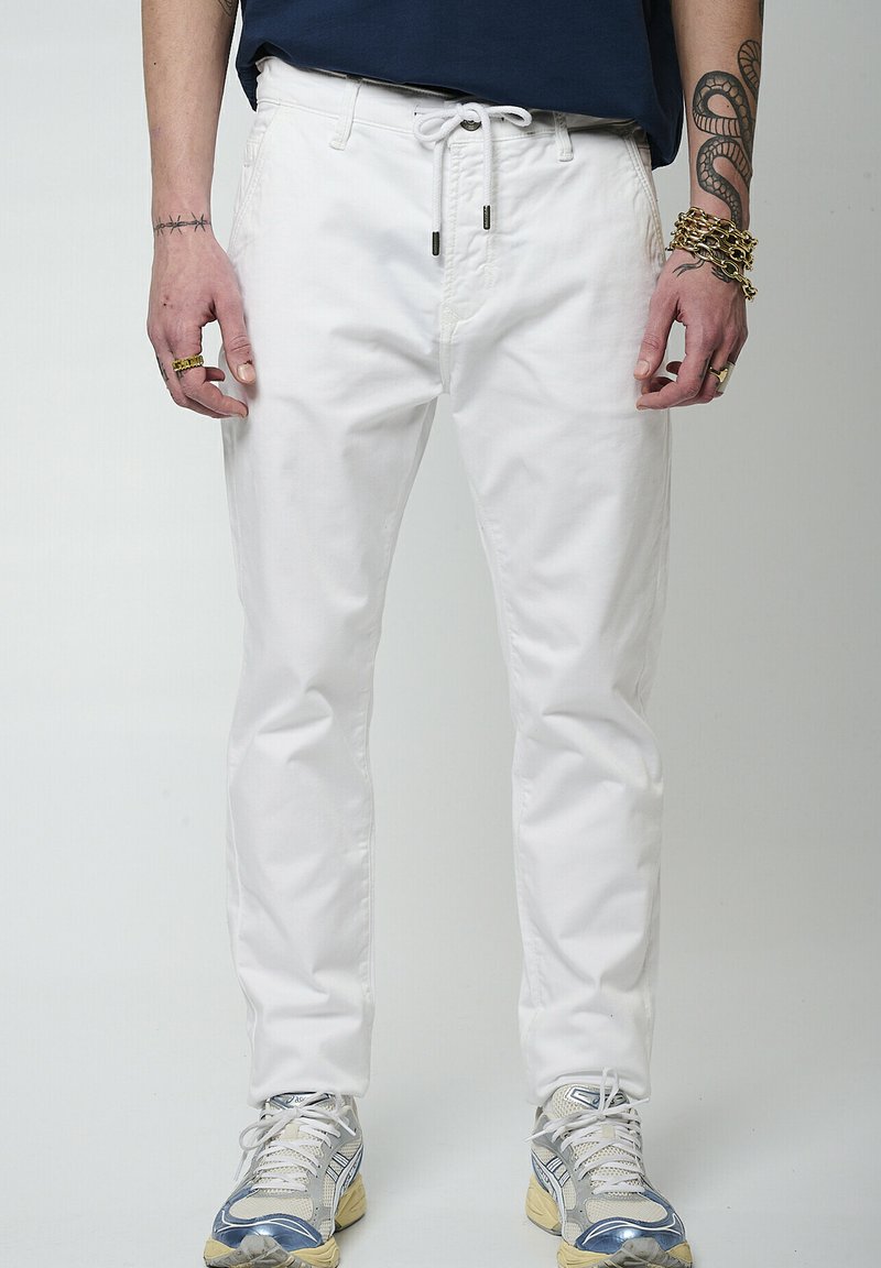 Pantalones de algodón blancos con un corte relajado, cintura con cordón y bolsillos laterales. Combinados con zapatillas grises y azules. Presenta intrincados tatuajes en la mano.