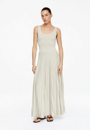 Kobieta ubrana w beżową, bezrękawnikową sukienkę maxi z plisowaną spódnicą, stoi z rozluźnionymi ramionami, ma na nogach czarne sandały na białym tle.