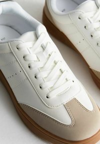 Vita lädersneakers med en beige mocka tåskydd, runda snörningar och en gummisula med bruna accenter. Minimalistisk design med perforeringar.