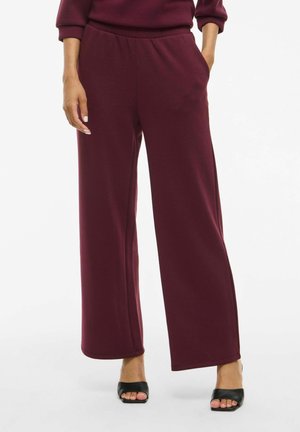 Pantalons larges bordeaux en tissu doux, avec une taille élastique et des poches latérales, associés à des sandales noires à bout ouvert.