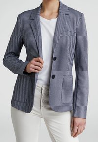 Blazer en tissu pied-de-poule texturé bleu marine et blanc, avec deux boutons, revers crantés et poches avant. Porté sur une chemise blanche.