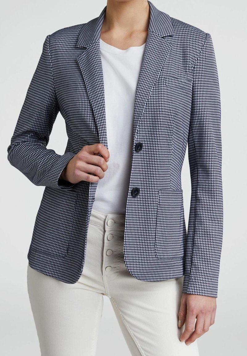 Blazer en tissu pied-de-poule texturé bleu marine et blanc, avec deux boutons, revers crantés et poches avant. Porté sur une chemise blanche.