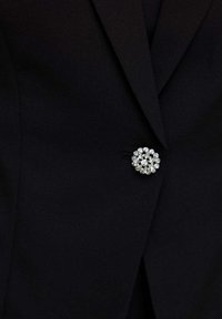 Blazer nero con tessuto testurizzato e un bottone rotondo e incastonato, con strass trasparenti al centro. Design della revers accentuato.