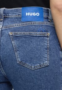Detailní záběr zadní kapsy modrých džínů s modrou značkou HUGO nahoře a nenápadným kruhovým logem vyšitým blízko kapsy.