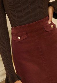Fustă de corduroy burgundy, cu buzunare frontale, butoane aurii și talie ajustată. Țesătura are un aspect texturat, cu caneluri.