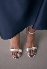 Sandales métalliques argentées avec deux larges brides. Le pied montre des ongles naturels. L'arrière-plan est d'une couleur neutre, réduisant le contraste.