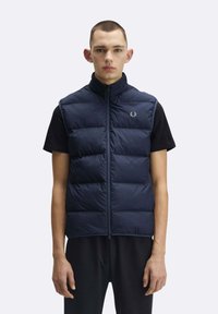 Giovane uomo che indossa un gilet trapuntato senza maniche blu navy sopra una maglietta nera a maniche corte, in piedi contro uno sfondo chiaro e uniforme.