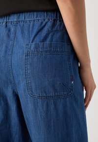 Tumši zilas denimu bikses ar elastīgu jostasvietu un aizmugurējo kabatu ar sarkaniem un baltiem šuvumiem. Gluda tekstūra.