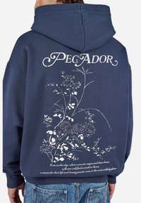 Marine hoodie met een wit bloemenontwerp en de tekst "PEGADOR" op de achterkant, gedragen door een persoon in blauwe jeans, met de rug naar de camera gekeerd.