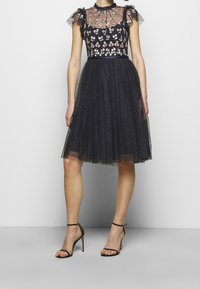 Robe noire avec broderie florale, superposition en tulle transparent, manches courtes et corsage ajusté, se déclinant en une jupe longueur genoux. Talons aiguilles noirs.