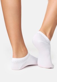 Calze bianche alla caviglia realizzate in un materiale liscio ed elastico con un motivo a zigzag lungo il bordo. Presentano un design a taglio basso.