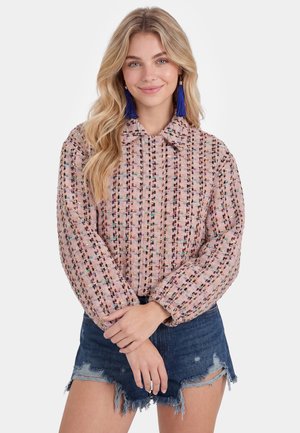 Junge Frau mit langen blonden Haaren trägt eine rosa gemusterte Jacke, blaue Quastenohrringe und ausgewaschene Denim-Shorts, lächelt mit verschränkten Händen.