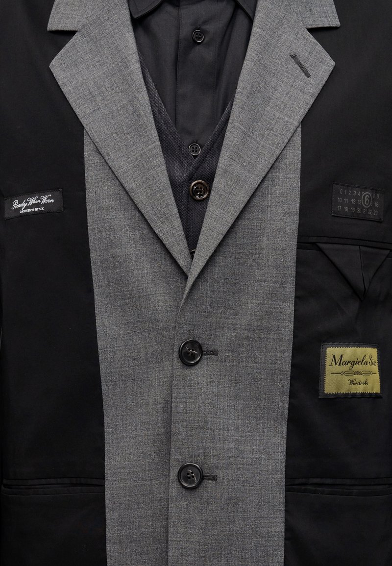 Blazer noir et gris avec un design bicolore, un revers gris texturé, cinq boutons, et plusieurs étiquettes sur la poche gauche.