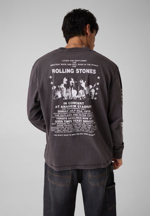 Uomo di spalle che indossa una maglia scura a maniche lunghe con stampa bianca della band Rolling Stones e testo riguardante un concerto del 1978 allo stadio di Anaheim.