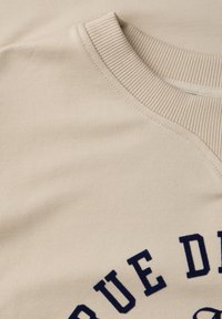 Sweat-shirt beige à col rond présentant des lettres en bloc bleu marine partiellement visibles, disposées en arc, sur un tissu doux.
