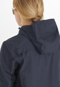 Marine lichtgewicht jack met een capuchon, voorzien van een gestructureerde stof en contrast zwarte accenten op de schouders en zijkanten.