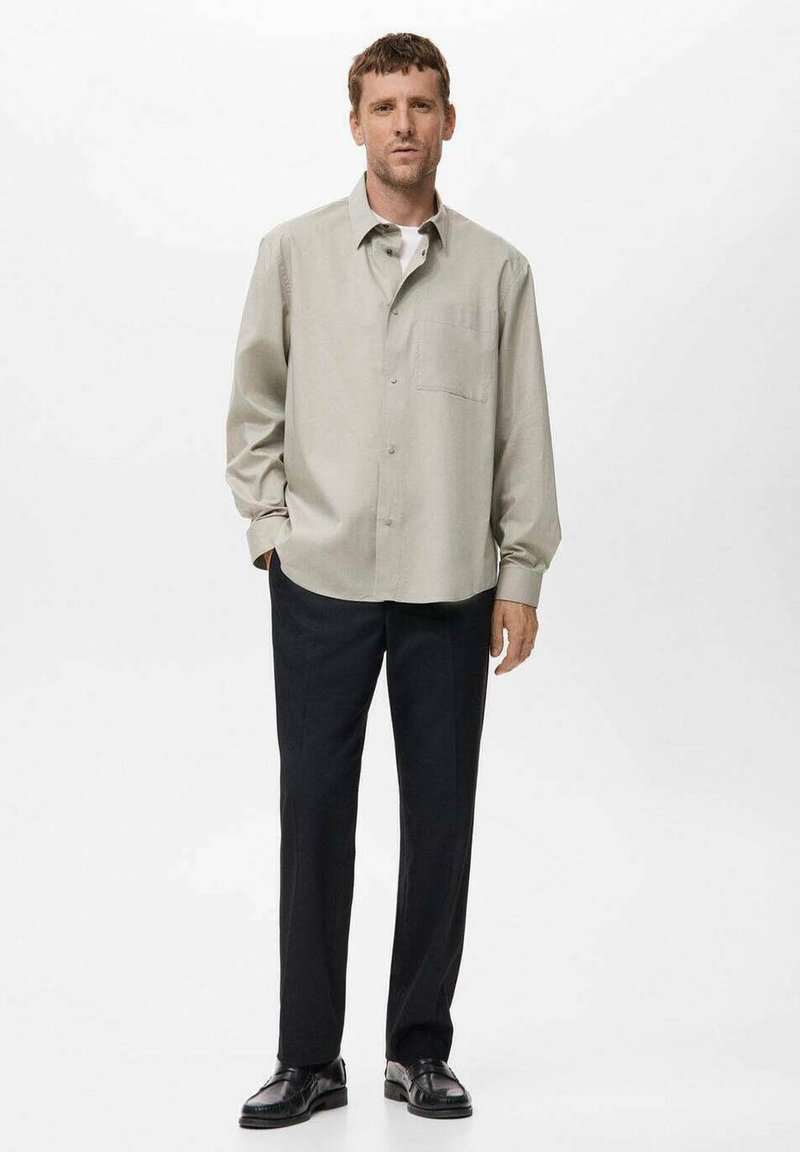 Mann steht bekleidet mit einem hellbeigen Hemd mit Knöpfen, schwarzen Hosen und schwarzen Anzugschuhen vor einem schlichten weißen Hintergrund.