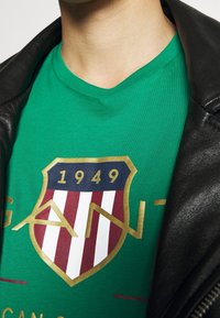 Zielona bawełniana koszulka z grafiką tarczy, w czerwono-białe paski, złotymi akcentami i rokiem "1949". Częściowo widoczna czarna skórzana kurtka.