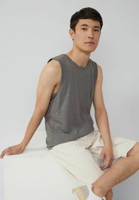 Graues ärmelloses Tanktop mit einer leicht strukturierten Oberfläche, kombiniert mit hellbeigen Shorts. Das Modell sitzt vor einem weißen Hintergrund.