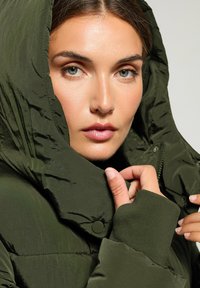 Giacca a vento imbottita di colore verde scuro con colletto alto, superficie texturizzata, bottoni a pressione e polsini elastici. Presenta una cerniera e un cappuccio.