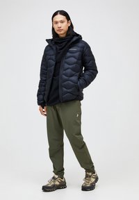 Black puffer jacket with a hood, featuring a quilted pattern. Worn over a black garment, paired with green pants and hiking shoes.