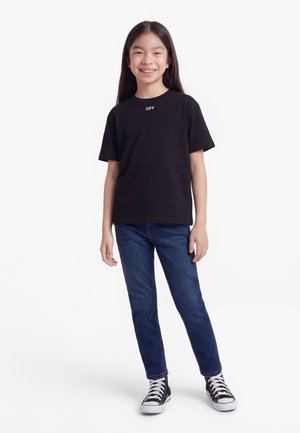 Νέος άνθρωπος που χαμογελά, φοράει μαύρο μπλουζάκι με το κείμενο "OFF", μπλε τζιν και μαύρα ψηλοτάκουνα παπούτσια, στέκεται μπροστά σε λευκό φόντο.