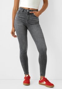 Pantalones vaqueros ajustados de tiro alto en gris con un ligero brillo, cinco bolsillos, trabillas para cinturón y cremallera, combinados con zapatillas rojas.