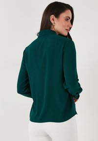 Dark green blouse with long sleeves, a collar, and button details at the cuffs. Smooth fabric with a relaxed fit and curved hemline.