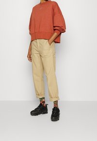 Persona che indossa un ampio felpa rossa, pantaloni beige con risvolto, calze a righe colorate con motivo floreale e scarpe da ginnastica nere, in piedi su uno sfondo bianco.