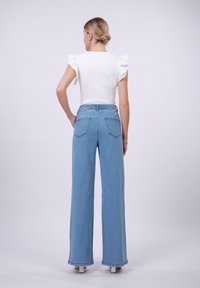 Svijetloplave široke traper hlače s stražnjim džepovima i opuštenim fitom, u kombinaciji s bijelom majicom s volanima na ramenima.