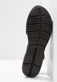 Semelle de chaussure en caoutchouc noir avec des grips texturés et des rainures, comportant des trous de ventilation et un logo. Surface lisse avec un design à bout arrondi.