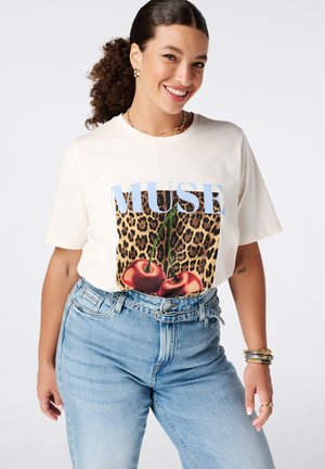 Vrouw glimlachend in een wit T-shirt met de tekst "MUSE", luipaardprint en kersgrafiek, gecombineerd met een high waist blauwe jeans en gouden sieraden.