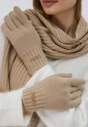 Guanti lavorati a maglia beige con polsini a coste e un'etichetta con il marchio. Texture morbida. Abbinati a una sciarpa beige coordinata. Tessuto accogliente, adatto per il freddo.