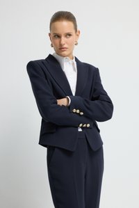 Granatowa marynarka o dopasowanym kroju z usztywnionymi ramionami, złotymi guzikami i gładką fakturą, zestawiona z białą koszulą z kołnierzykiem i dopasowanymi spodniami.