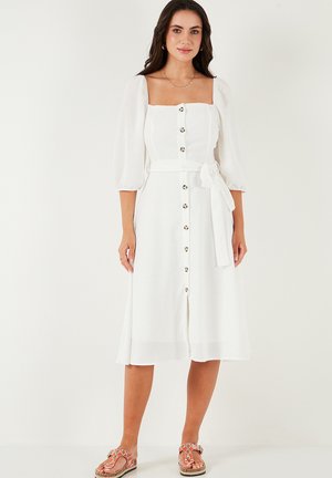 White linen dress with a square neckline, puffed sleeves, front button closure, and a waist tie. Features a flowy midi length and subtle texture.