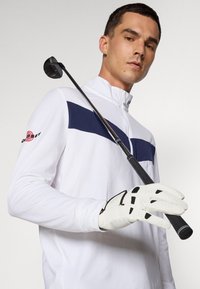 Valge pika varrukaga golfisärk, millel on tumesinine horisontaalne triip, koos valge golfikindaga ja hoides golfiklubiga.