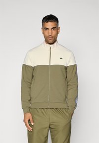 Olijfgroene trainingsjack met een crèmekleurig bovengedeelte, ritsluiting, hoge kraag en blauwe accenten langs de mouwen. Bevat het Lacoste-logo.