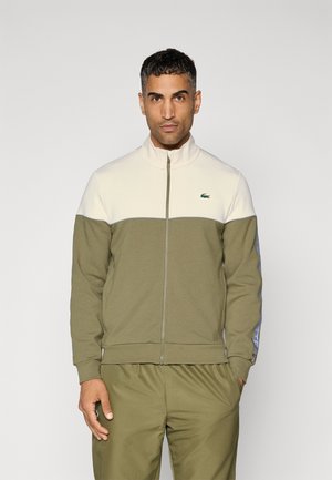 Giacca di tracksuit verde oliva con una parte superiore crema, chiusura con zip, colletto alto e dettagli blu lungo le maniche. Presenta il logo Lacoste.