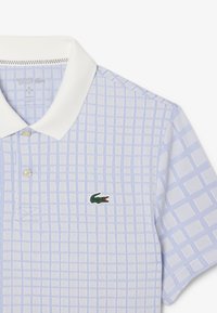 Ljusblå pikétröja med vit krage, med ett rutmönster och en grön broderad krokodil-logotyp på bröstet. Texturerad tyg.